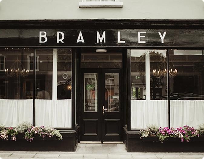 The front of Preston House café named BRAMLEY features a black exterior, white curtains in the windows, and flower boxes filled with pink and white flowers along the bottom of the windows.