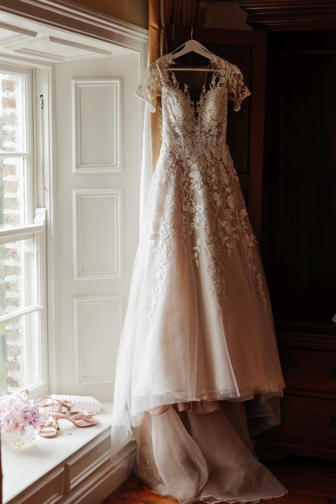 A white lace wedding dress with floral details hangs near a window at Preston House. Pink shoes and a floral hair accessory rest on the windowsill, whilst warm natural light illuminates the elegant scene.
