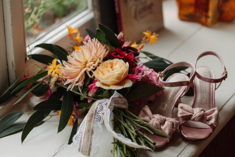 A bouquet of assorted flowers with pink, yellow, and orange blooms rests on a Preston House windowsill next to a pair of pink heeled sandals with bows, whilst a white lace ribbon drapes from the bouquet.