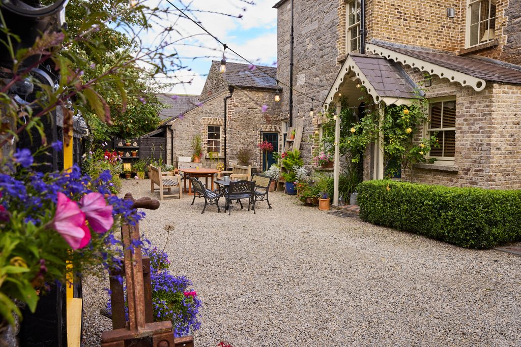 A gravel courtyard at Preston House with outdoor tables and chairs, surrounded by stone buildings, lush greenery, colourful flowers, and fairy lights overhead, creates a cosy, inviting garden setting.