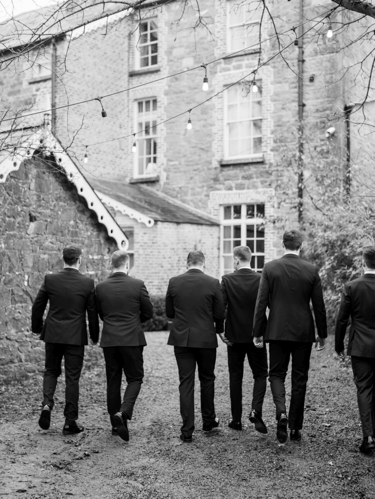 Six men in suits walk away from the camera down a gravel path beside stone buildings at Preston House, with fairy lights overhead and bare tree branches visible in this black and white photo.