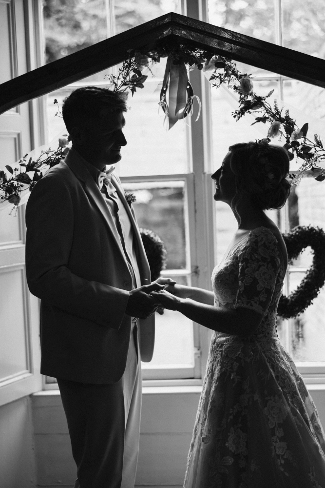 A bride and groom stand facing each other, holding hands under a floral arch by a bright window at Preston House. Their silhouettes suggest they are sharing a romantic, intimate moment indoors.