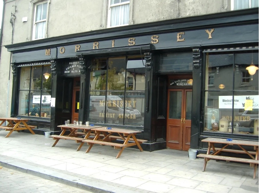 A traditional pub with a black exterior, gold “MORRISSEY” sign above large windows, and wooden doors. Wooden picnic tables sit on the pavement outside Preston House, inviting guests to relax and unwind.