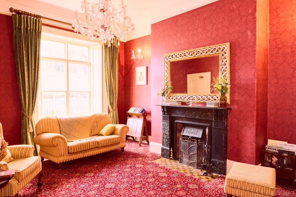 A bright, elegant living room in Preston House features red patterned wallpaper and carpet, a large window with green curtains, yellow upholstered sofas, a dark ornate fireplace, a decorative mirror, and a chandelier hanging from the ceiling.