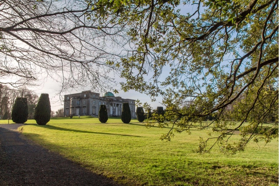 The historic Preston House, a large stone building with columns, stands on a green lawn surrounded by manicured trees under a clear blue sky. Tree branches with green leaves frame the top of the image.