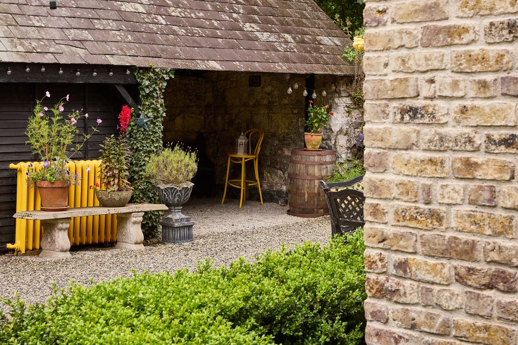 A rustic garden scene at Preston House with a stone bench, potted flowers, a yellow radiator, a wooden barrel, and a single yellow chair under a slanted roof beside a brick wall and green bushes.