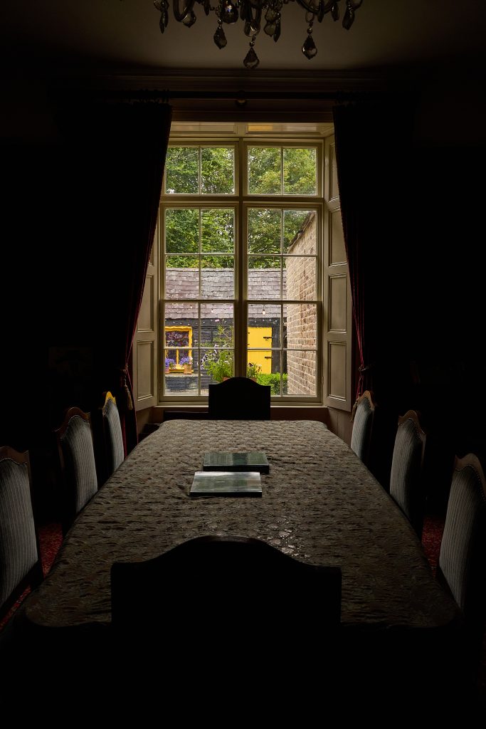 A dimly lit dining room with a long table and chairs faces a large window at Preston House. Outside, a yellow building and lush greenery are visible, illuminated by daylight.
