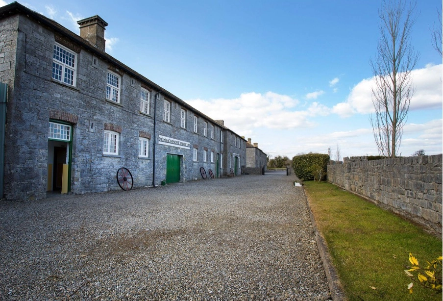 Preston House, a long stone building with green doors and arched windows, stands beside a gravelled courtyard bordered by a low stone wall and a small patch of grass under a partly cloudy sky.