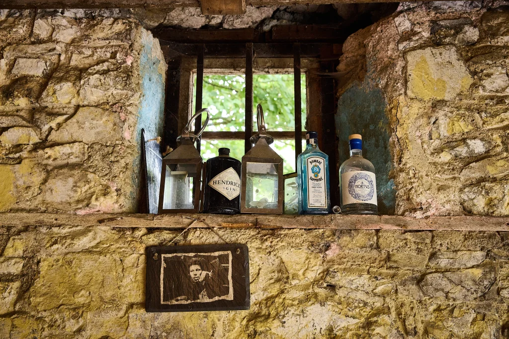 At Preston House, a rustic stone windowsill holds two metal lanterns and three bottles of gin beneath a barred window. Below the sill, a sepia photo of a man in a frame is mounted on the wall. Green foliage is visible outside.