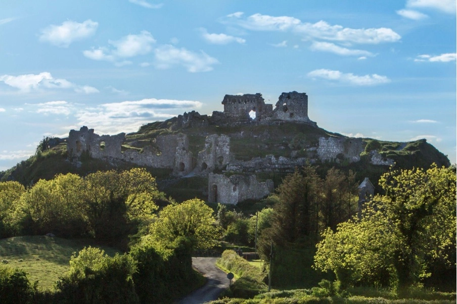 Ruins of an ancient stone castle sit atop a grassy hill near Preston House, surrounded by green trees and bushes under a blue sky with scattered clouds. A narrow road winds through the foreground.