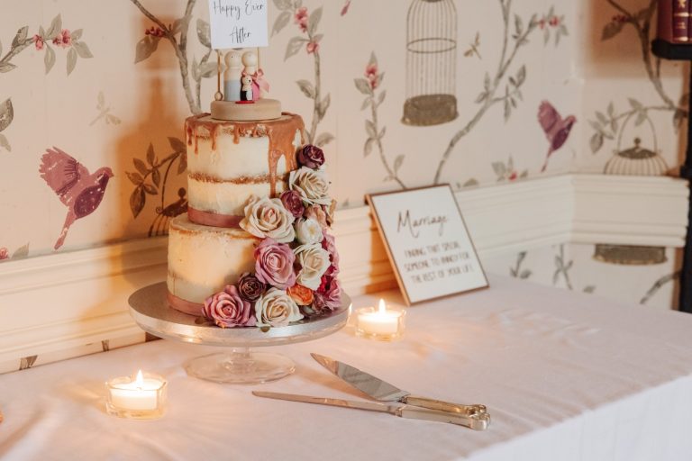 A two-tier wedding cake decorated with pink and white roses and a small figurine topper sits on a table at Preston House, beside lit candles, a cake knife, and a framed marriage sign, against a floral wallpapered wall.