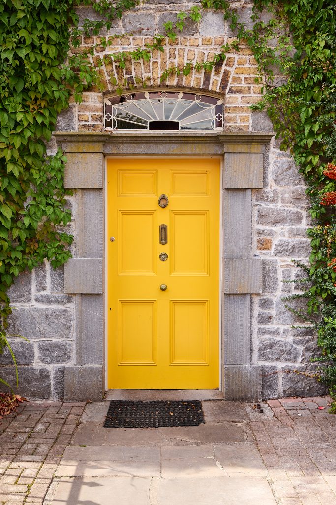 A bright yellow door with panel detailing welcomes you to Preston House, set in a grey stone wall and framed by climbing green ivy. A decorative fanlight window tops the entrance, whilst a small black doormat lies in front.