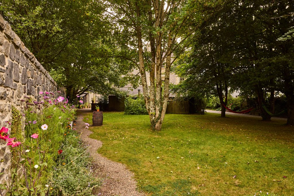 A lush garden at Preston House features tall trees, a stone wall on the left lined with colourful flowers, a gravel path curving through grass, and a hammock hanging between trees in the background near a building.