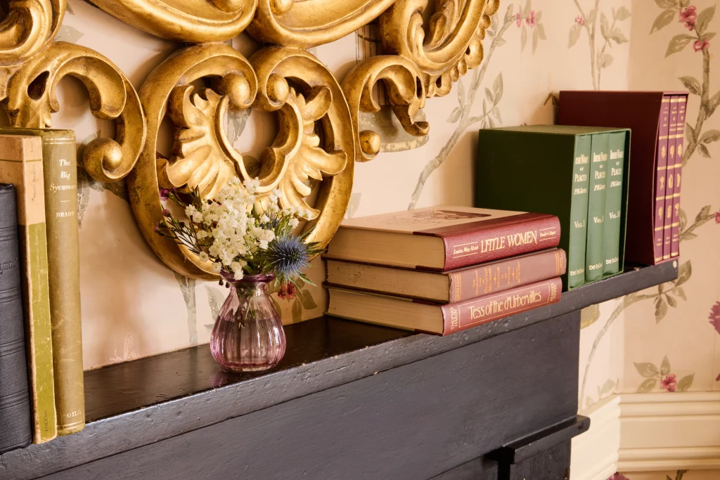 A black mantelpiece in Preston House holds a small glass vase with white flowers and thistles, several large green and red books, and three stacked classic novels, with ornate gold decor and floral wallpaper in the background.