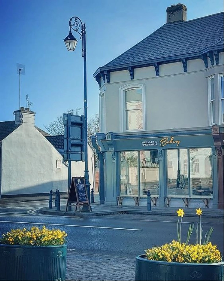 Street view of Muller & O’Connell Bakery near Preston House on a sunny day, with yellow daffodils in planters in the foreground and a streetlamp and chalkboard sign by the bakery entrance.