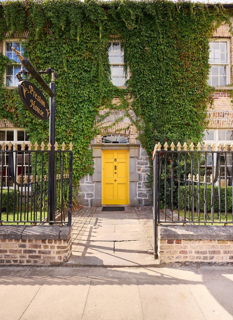 Preston House stands as a brick building draped in green ivy, featuring a bright yellow door, black wrought iron fence, and a hanging sign. Two windows overlook the entrance of this charming property.