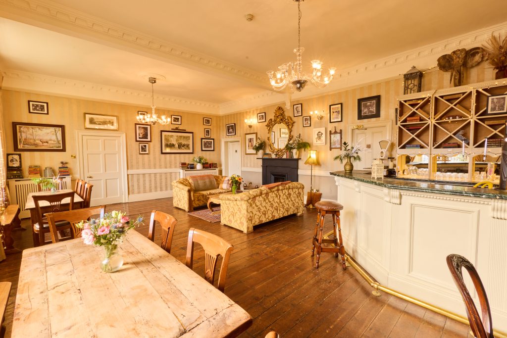 Preston House features a cosy vintage-style lounge with wooden floors, chandeliers, floral sofas, a rustic bar, and framed pictures. Fresh flowers on wooden tables complete the warm, inviting atmosphere.