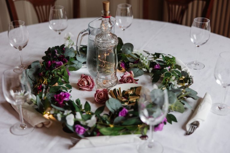 A round table at Preston House is set with empty wine glasses, a clear glass water jug and bottle, and a centrepiece of green foliage, purple and pink flowers, and fairy lights on a white tablecloth.