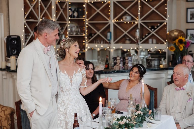 A bride in a lace dress gestures whilst speaking beside the groom in a cream suit at Preston House. Guests sit at a table with candles and drinks, while shelves with bottles and fairy lights glow in the background.