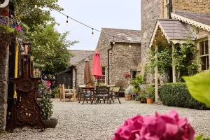A charming courtyard at Preston House with gravel ground, outdoor dining tables and chairs, colourful umbrellas, fairy lights, potted plants, flowers, and stone buildings with ivy and wooden accents on a clear day.
