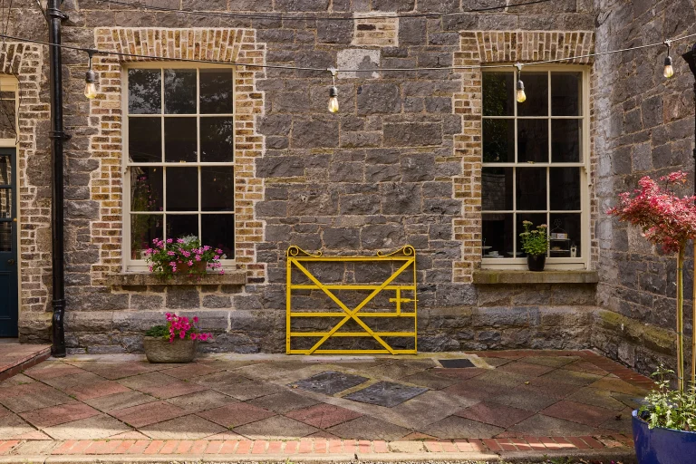 A brick courtyard at Preston House features two large windows, potted flowers, a hanging string of lights, and a bright yellow metal gate against the stone wall. The ground is paved with patterned stone tiles.