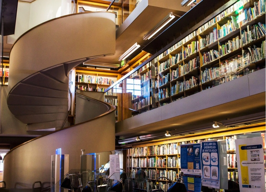 A modern library interior at Preston House features tall bookshelves, glass panels, and a large spiral staircase. Posters and chairs are visible in the foreground, whilst bright lights illuminate the inviting space.