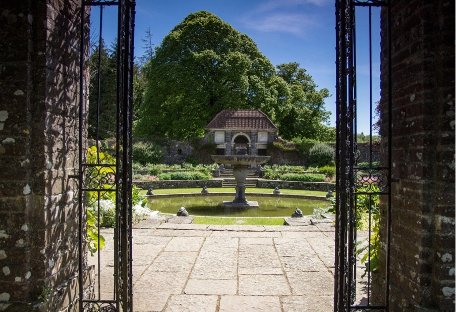 View through iron gates of a stone path leading to a circular pond with central fountain, surrounded by garden beds and greenery at Preston House; a stone building and large tree stand under a blue sky.