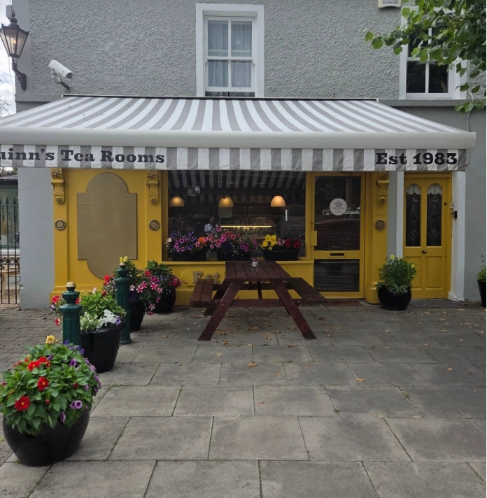 A quaint tearoom, Preston House, with a striped awning, yellow trim, and flower-filled planters outside. A wooden picnic table sits on the pavement in front. The sign reads Quinns Tea Rooms Est 1983.