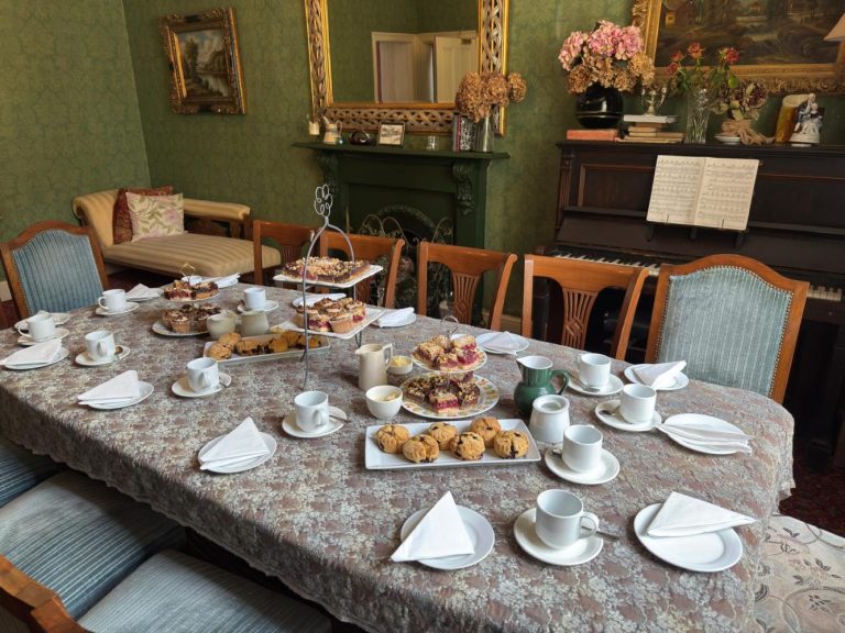 A dining table at Preston House set for afternoon tea with cups, saucers, serviettes, and plates of pastries and scones. The room features vintage decor, a piano with sheet music, a fireplace, and framed art on the walls.