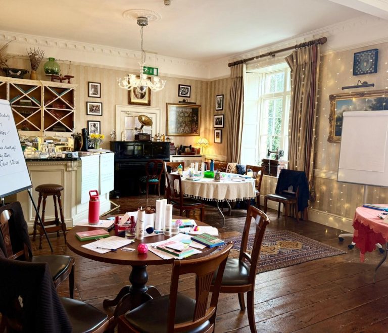 A cosy, well-lit room at Preston House set up for a workshop, with round tables, chairs, assorted papers and stationery, a flip chart, bookshelves, framed pictures on the walls, and a large window letting in natural light.
