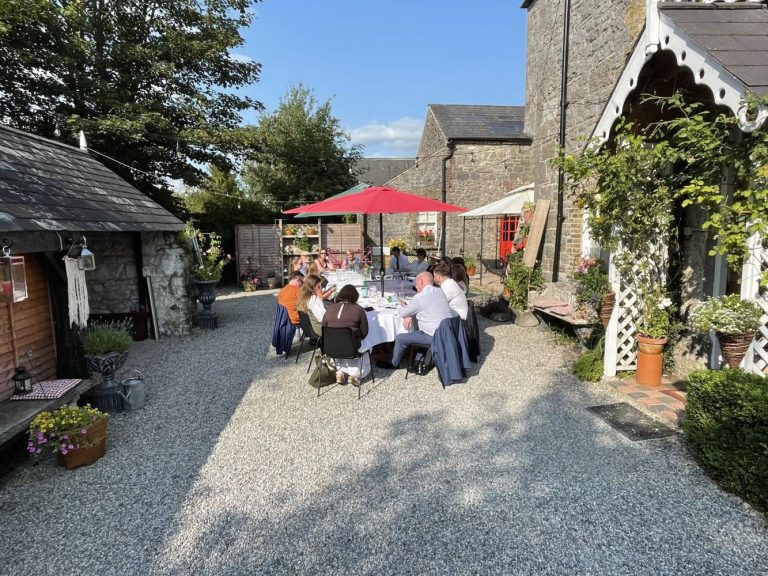 A group of people sit around a table under a red umbrella in the sunny stone courtyard of Preston House, surrounded by plants and rustic buildings, sharing a meal and conversation.