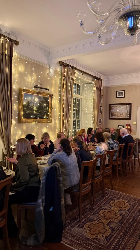A group of people sit around a long wooden table in the elegant dining room of Preston House, decorated with fairy lights, paintings, and large windows with curtains, enjoying a meal together.