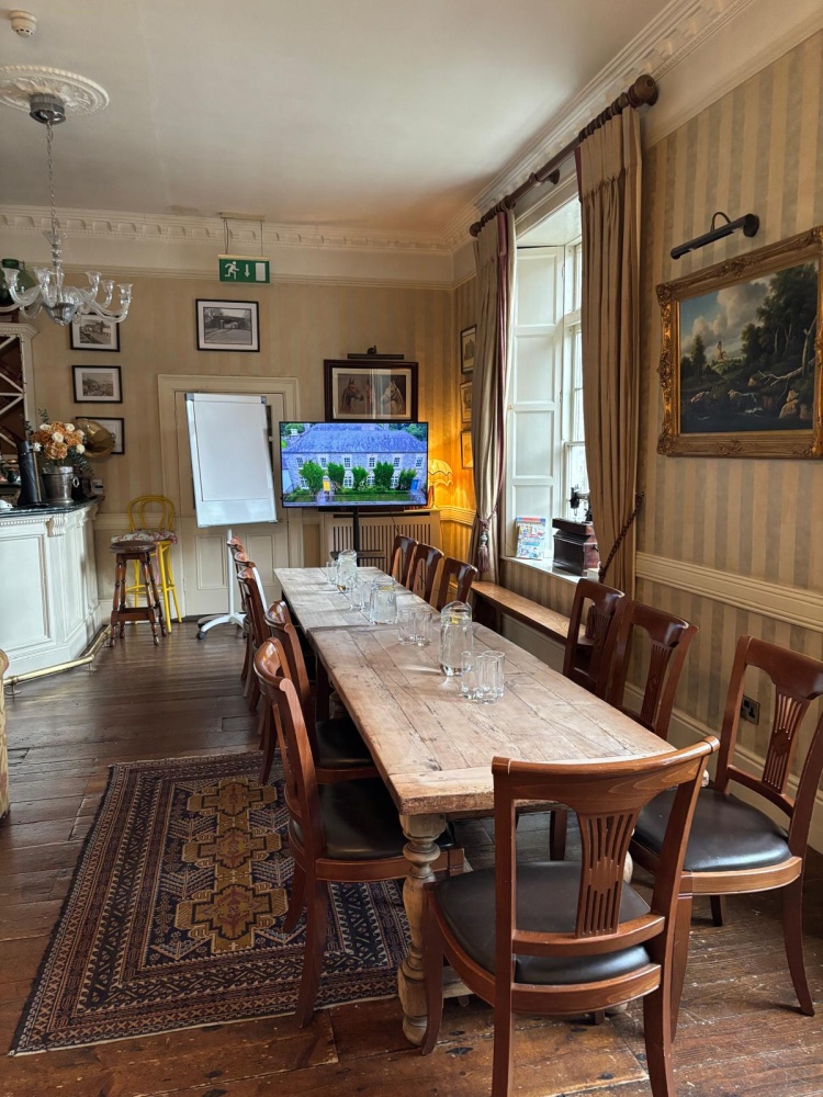 A cosy dining room at Preston House features a long wooden table set for eight with clear glasses, wooden chairs, a patterned rug, paintings on striped walls, a flipchart, and a TV beside a large window letting in natural light.