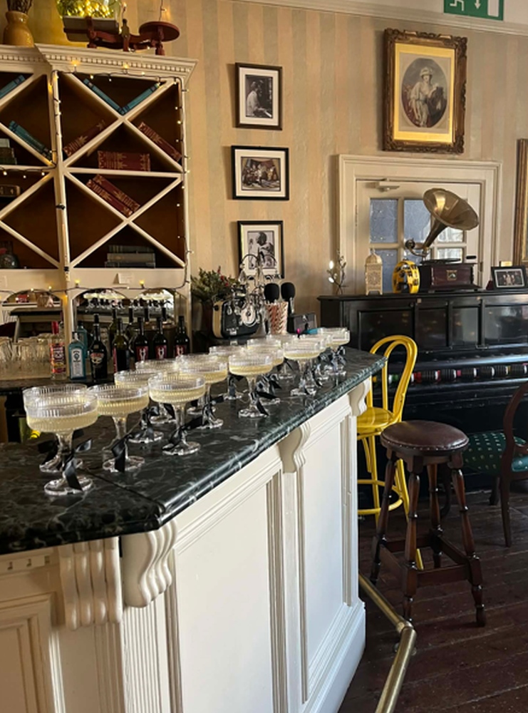 A vintage-style bar at Preston House, with glasses lined up on a marble counter, a piano, bottles, framed photos on the wall, a gramophone, and a yellow chair in a cosy, well-lit setting.