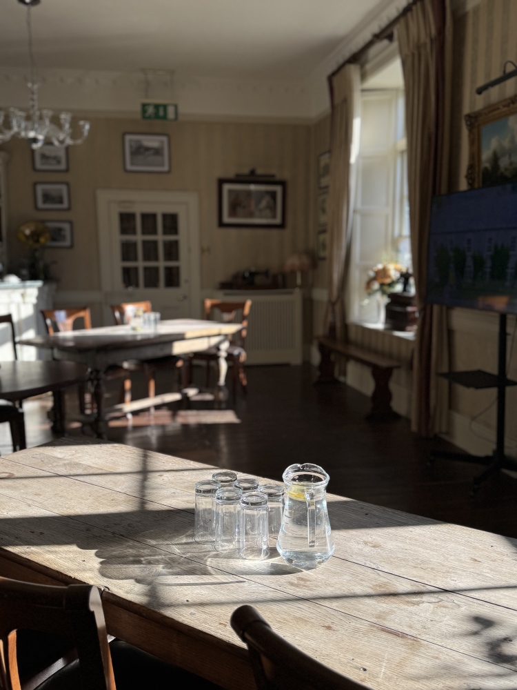 A sunlit room at Preston House with wooden tables and chairs, featuring a glass jug of water with a slice of lemon and four upturned glasses on a table in the foreground. Large windows let in natural light.