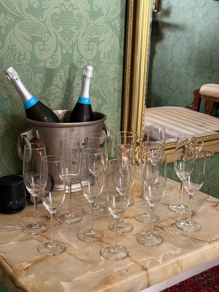 Two bottles of champagne in an ice bucket sit on a marble table at Preston House, surrounded by empty champagne flutes. The scene is set against green patterned wallpaper and a large gold-framed mirror.
