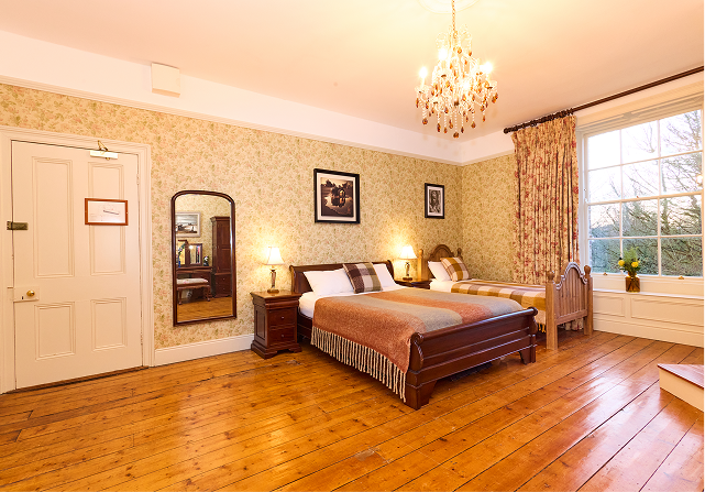 A spacious bedroom in Preston House with wooden floors, a double bed and a single bed, vintage wallpaper, framed art on the walls, a chandelier, a large window with curtains, and bedside tables with lamps.