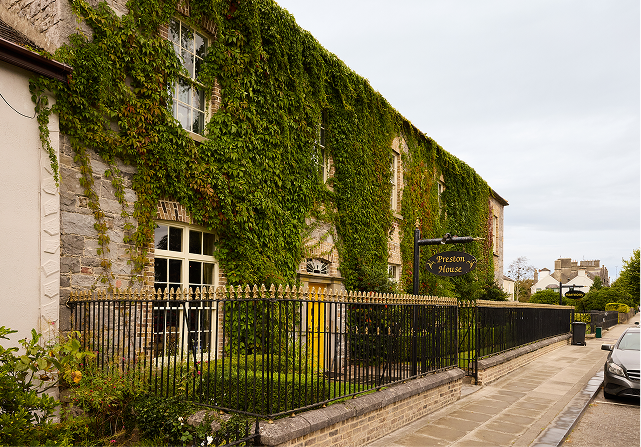 A stone building covered in green ivy stands behind a black iron fence on a quiet, empty street. A sign reading “Preston House” marks its entrance, whilst parked cars and other buildings line the pavement beyond.