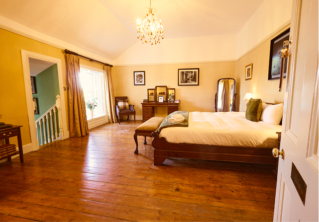 Spacious, warmly lit Preston House bedroom with wooden floor, a large bed with white linen, chandelier, wooden furniture, framed pictures on the walls, and a window with beige curtains letting in natural light.