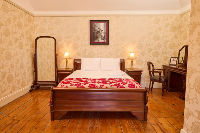 A cosy Preston House bedroom with a wooden bed, white bedding, and a red patterned throw, flanked by bedside tables with lamps, a standing mirror, a desk with a chair, and ornate wallpaper.