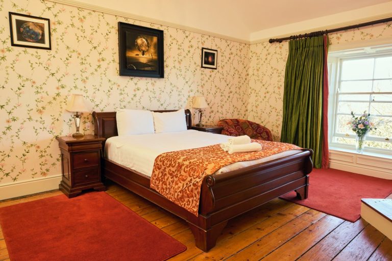 A cosy Preston House bedroom with a wooden bed, white linen, and an orange patterned throw. Two bedside tables with lamps, floral wallpaper, red rugs, green curtains, a window, and framed art complete the inviting space.