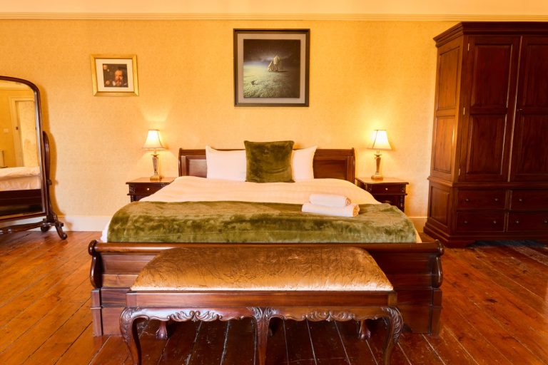 A cosy Preston House bedroom with a large wooden bed, green and white bedding, two bedside lamps, a wooden bench at the foot of the bed, a wardrobe, mirror, and framed pictures on yellow patterned walls.
