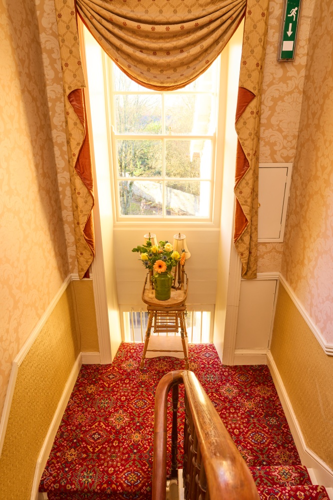 A bright stairwell in Preston House features ornate carpet, patterned wallpaper, a large window with gold curtains, and a small table holding a vase of yellow flowers beside a lamp. An exit sign is mounted on the wall above.