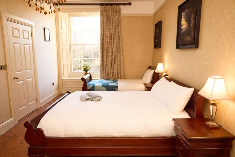 A cosy Preston House bedroom with a large bed and a single bed, wooden furniture, two bedside lamps, cream curtains, a vase with flowers, and framed art. Warm lighting creates a welcoming atmosphere.