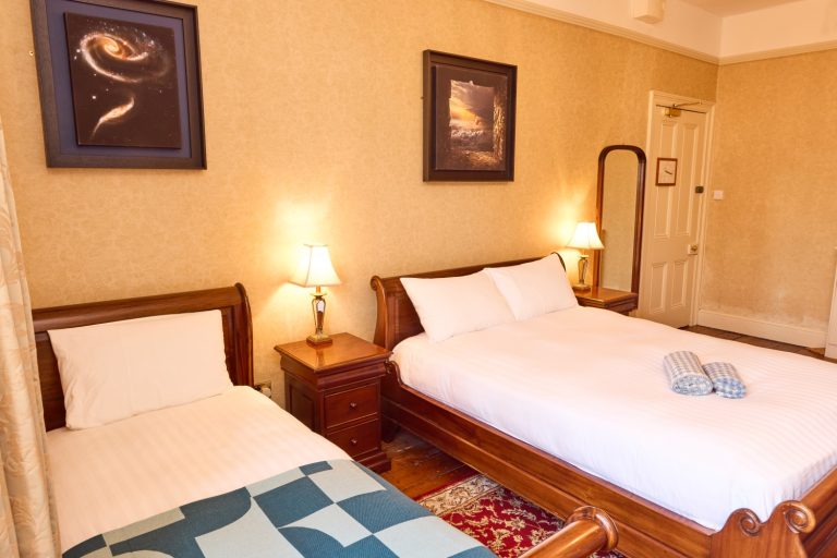 A cosy Preston House hotel room with one single and one double wooden bed, white bedding, two wall lamps, framed art on beige walls, a tall mirror, and neatly folded towels on the double bed.