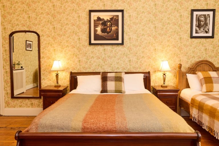 A cosy Preston House bedroom with a double bed and a single bed, both draped in checked blankets. Two bedside tables with lamps, floral wallpaper, framed photos on the wall, and a mirror reflect part of the room.