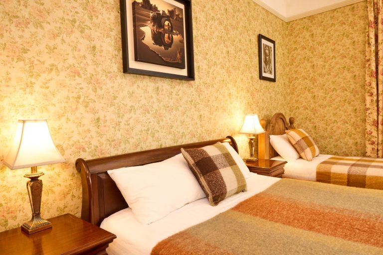 A cosy bedroom at Preston House features a double bed and a single bed, both with checked blankets and pillows. Two bedside tables with lamps and patterned wallpaper add a warm, vintage atmosphere, whilst framed pictures hang on the wall.