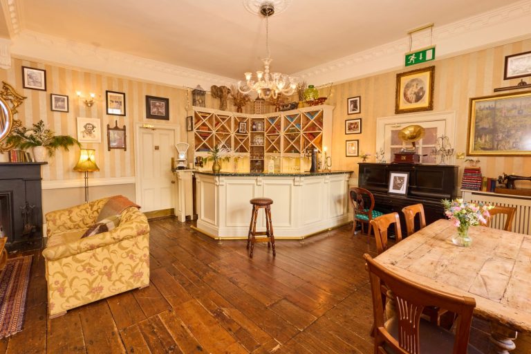 A cosy vintage-style room in Preston House with wooden floors, a cream-coloured bar, chandelier, framed pictures on striped walls, an upholstered sofa, wooden dining table with flowers, and antique décor elements.