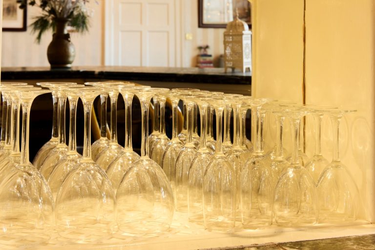Rows of upside-down wine glasses are neatly arranged on a counter at Preston House, with a blurred background featuring a vase with flowers and interior decor.