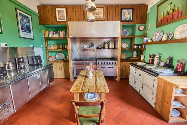 A cosy, vintage kitchen in Preston House features green walls, wooden shelves with dishes and cookery books, a large cooker, and a wooden table with a single chair. Decorative plates and framed art complete the inviting space.